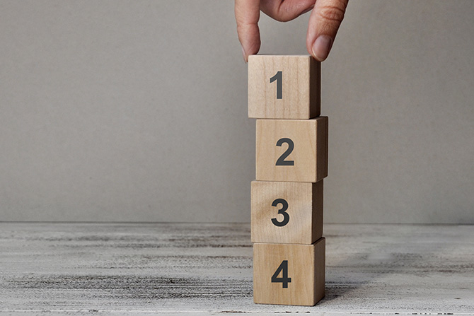 From the Top: Setting Our Priorities for 2026; A hand stacks wooden blocks numbered 1 to 4 in a vertical tower on a textured surface, symbolizing growth and progression in a neutral setting.