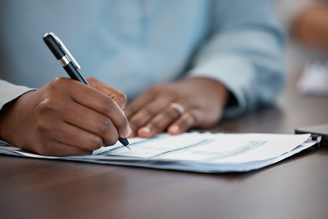 Industrial Loan Bank Charters; A person writes with a pen on documents at a table, their focus evident. The setting suggests a professional environment, conveying concentration and diligence.